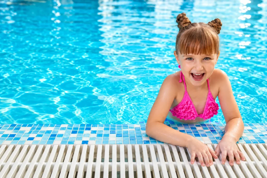 Petite fille souriante en maillot rose, dans l’eau, les mains posées sur le bord de la piscine.